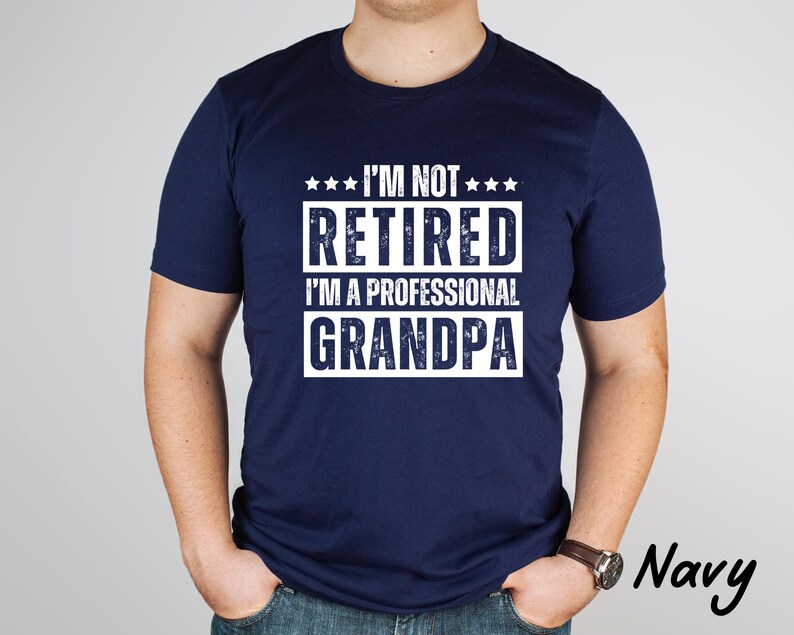 May include: Navy blue t-shirt with the text "I'M NOT RETIRED I'M A PROFESSIONAL GRANDPA" in white. The shirt has a crew neck and short sleeves. The word "Navy" is printed in the bottom right corner.