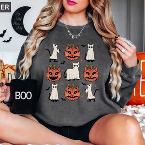 K&ouml;nnte beinhalten: Dunkelgraues Sweatshirt mit einem Halloween-Design. Das Design zeigt Cartoon-Katzen, K&uuml;rbisse und Geister. Eine schwarze Tasse mit dem Wort "BOO" ist zu sehen. Das Wort "PEPPER" befindet sich oben links.
