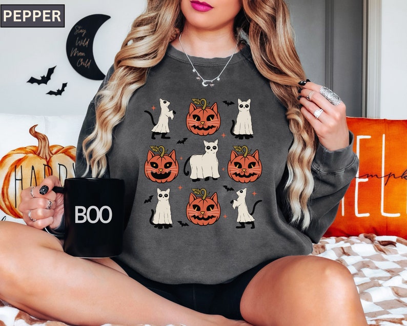 K&ouml;nnte beinhalten: Dunkelgraues Sweatshirt mit einem Halloween-Design. Das Design zeigt Cartoon-Katzen, K&uuml;rbisse und Geister. Eine schwarze Tasse mit dem Wort "BOO" ist zu sehen. Das Wort "PEPPER" befindet sich oben links.