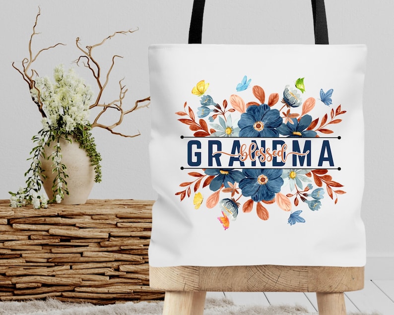 K&ouml;nnte beinhalten: Wei&szlig;e Einkaufstasche mit einem Blumendesign in Blaut&ouml;nen, Orange und Gr&uuml;n. Der Text "blessed grandma" ist in Blau auf die Tasche gedruckt.