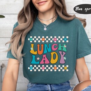 May include: A teal blue t-shirt with a checkered pattern and the text "Lunch Lady" in colorful retro letters. The letters are outlined in white and have small flowers on them.