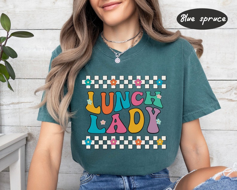 May include: A teal blue t-shirt with a checkered pattern and the text "Lunch Lady" in colorful retro letters. The letters are outlined in white and have small flowers on them.