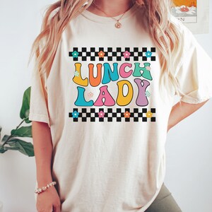 May include: A white t-shirt with a checkered border and the text "Lunch Lady" in colorful, retro-style letters. The letters are outlined in black and have small flowers on them.