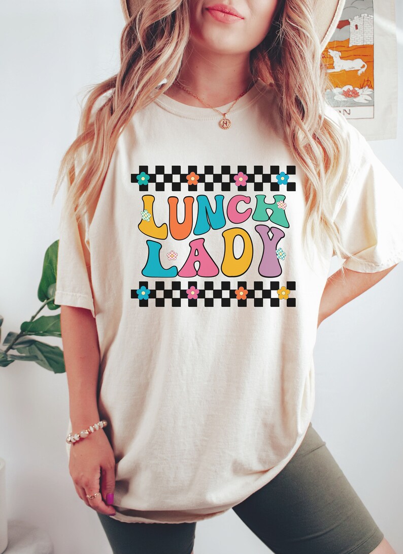 May include: A white t-shirt with a checkered border and the text "Lunch Lady" in colorful, retro-style letters. The letters are outlined in black and have small flowers on them.
