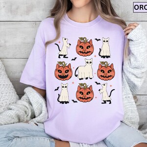 K&ouml;nnte beinhalten: Ein helllila T-Shirt mit einem sich wiederholenden Halloween-Muster. Das Design umfasst orangefarbene K&uuml;rbislaternen, wei&szlig;e Geisterfiguren, schwarze Katzen und Flederm&auml;use. Das Wort "ORCHID" ist in einem wei&szlig;en Rechteck oben rechts aufgedruckt.