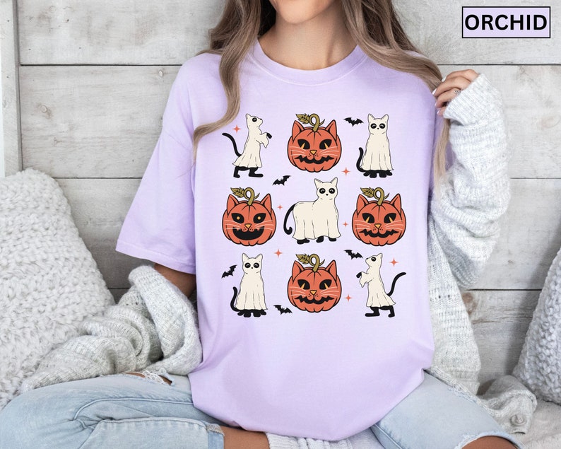 K&ouml;nnte beinhalten: Ein helllila T-Shirt mit einem sich wiederholenden Halloween-Muster. Das Design umfasst orangefarbene K&uuml;rbislaternen, wei&szlig;e Geisterfiguren, schwarze Katzen und Flederm&auml;use. Das Wort "ORCHID" ist in einem wei&szlig;en Rechteck oben rechts aufgedruckt.