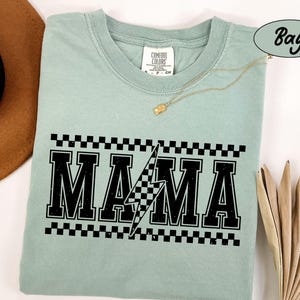 May include: A light green t-shirt with the word "MAMA" in bold black letters, a lightning bolt, and a checkered pattern. The shirt has a crew neck and short sleeves. A gold necklace is draped across the shirt.
