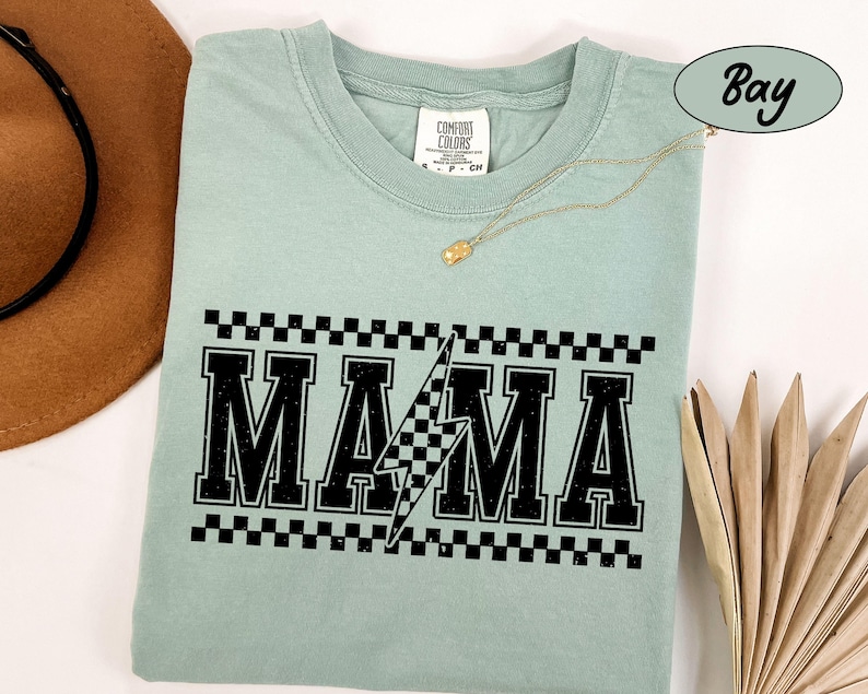 May include: A light green t-shirt with the word "MAMA" in bold black letters, a lightning bolt, and a checkered pattern. The shirt has a crew neck and short sleeves. A gold necklace is draped across the shirt.