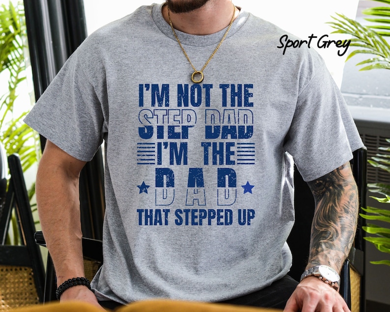 Puede incluir: Una camiseta gris con el texto "I'm not the step dad I'm the dad that stepped up" en letras azules. La camiseta tiene un aspecto desgastado y presenta una estrella a cada lado de la palabra "dad".