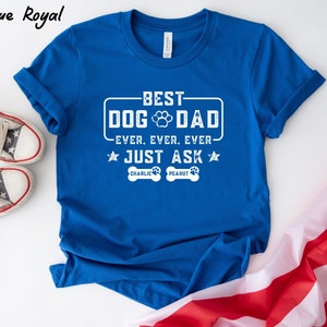 May include: A royal blue t-shirt with white text that reads "BEST DOG DAD EVER. EVER. EVER. JUST ASK." The shirt includes a paw print graphic and the names "CHARLIE" and "PEANUT" on dog bone shapes. The shirt is folded, with a pair of star-spangled sneakers and a flag in the background.