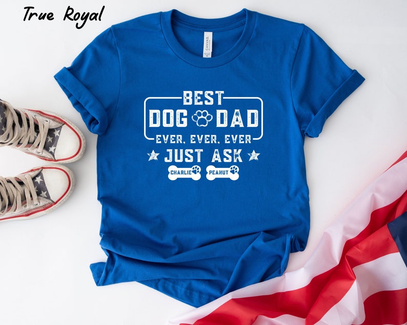 May include: A royal blue t-shirt with white text that reads "BEST DOG DAD EVER. EVER. EVER. JUST ASK." The shirt includes a paw print graphic and the names "CHARLIE" and "PEANUT" on dog bone shapes. The shirt is folded, with a pair of star-spangled sneakers and a flag in the background.