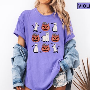 K&ouml;nnte beinhalten: Violettes T-Shirt mit Halloween-Motiv. Das Design zeigt Cartoon-K&uuml;rbisse mit Katzen-Gesichtern, wei&szlig;e Geister und schwarze Flederm&auml;use. Das Wort "VIOLET" befindet sich in einem Feld oben rechts.