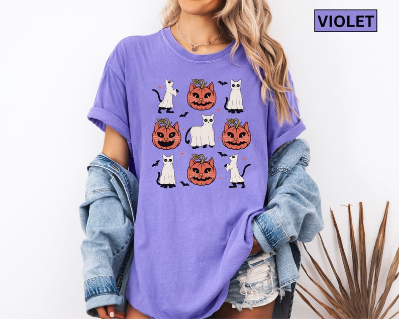 K&ouml;nnte beinhalten: Violettes T-Shirt mit Halloween-Motiv. Das Design zeigt Cartoon-K&uuml;rbisse mit Katzen-Gesichtern, wei&szlig;e Geister und schwarze Flederm&auml;use. Das Wort "VIOLET" befindet sich in einem Feld oben rechts.