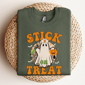 May include: An olive green sweatshirt with the words "STICK OR TREAT" in orange. The design features a cartoon ghost in a nurse's hat, holding a syringe and a jack-o'-lantern, with bats, a spider web, and a checkered background.