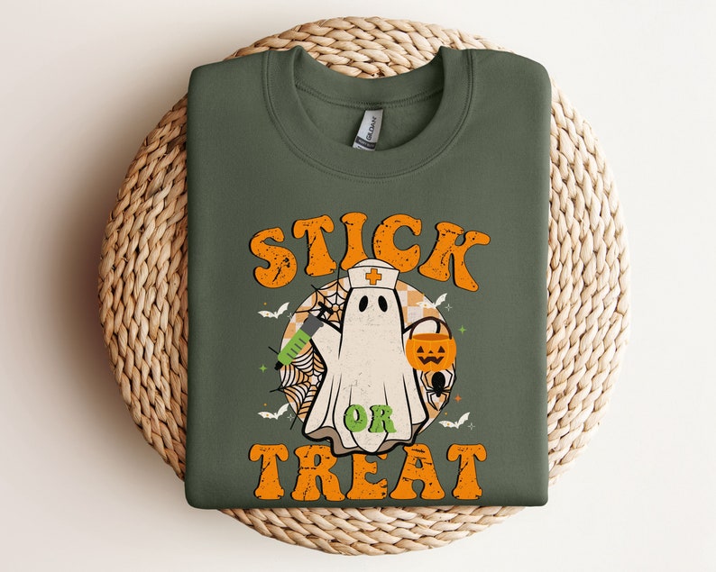 May include: An olive green sweatshirt with the words "STICK OR TREAT" in orange. The design features a cartoon ghost in a nurse's hat, holding a syringe and a jack-o'-lantern, with bats, a spider web, and a checkered background.