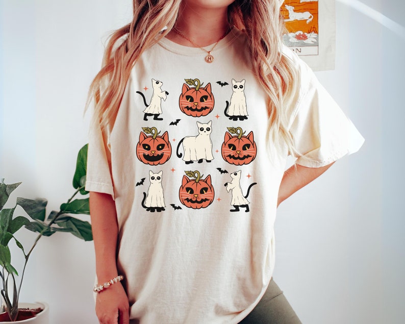 K&ouml;nnte beinhalten: Cremefarbenes T-Shirt mit einem Raster von Halloween-Illustrationen. Das Design umfasst Cartoon-Geister, K&uuml;rbislaternen und schwarze Katzen. Die K&uuml;rbisse sind orange mit gr&uuml;nen Stielen, und die Katzen und Geister sind wei&szlig;.