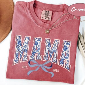 Może przedstawiać: Czerwony t-shirt w kolorze karmazynowym z białym i niebieskim nadrukiem kwiatowym, na kt&oacute;rym widnieje napis "MAMA" z niebieską kokardką i tekstem "EST. 2025" poniżej.