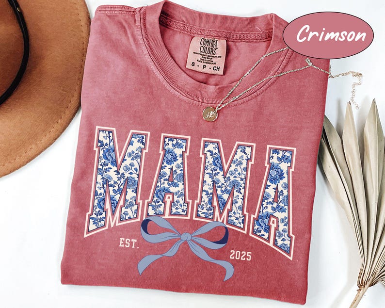 Może przedstawiać: Czerwony t-shirt w kolorze karmazynowym z białym i niebieskim nadrukiem kwiatowym, na kt&oacute;rym widnieje napis "MAMA" z niebieską kokardką i tekstem "EST. 2025" poniżej.