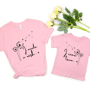 Op de afbeelding: Twee roze T-shirts met paardenbloemontwerpen en tekst. Het grotere shirt zegt "Ik heb een wens gedaan" en het kleinere shirt zegt "Ik ben uitgekomen".