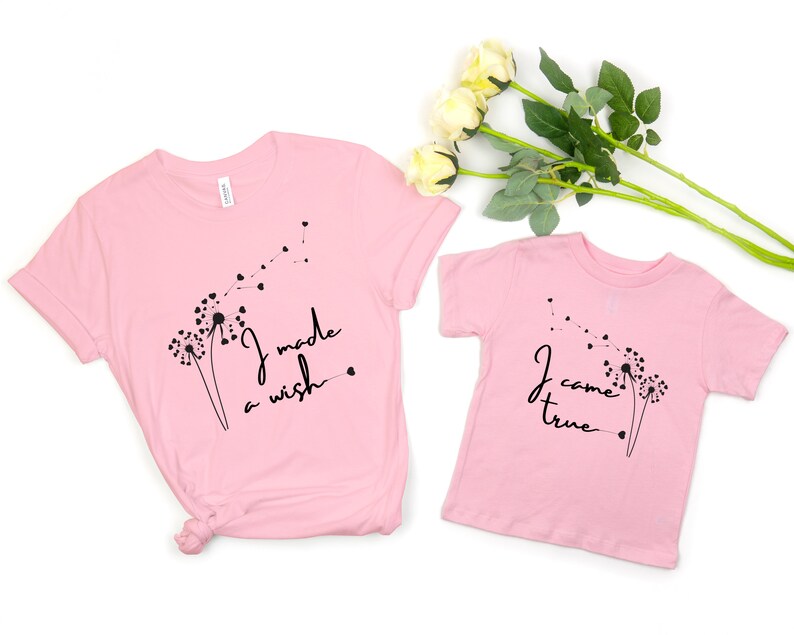 Op de afbeelding: Twee roze T-shirts met paardenbloemontwerpen en tekst. Het grotere shirt zegt "Ik heb een wens gedaan" en het kleinere shirt zegt "Ik ben uitgekomen".