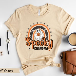 May include: A soft cream-colored t-shirt with a Halloween-themed graphic. The design features a ghost holding a pumpkin, a rainbow, spiders, and the words "Spooky Grandma" in orange. The shirt is made of a soft material.