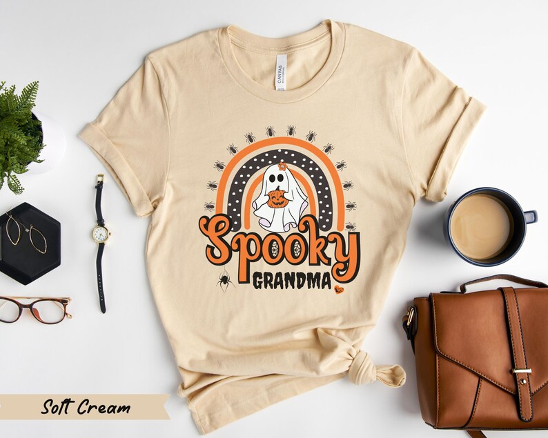 May include: A soft cream-colored t-shirt with a Halloween-themed graphic. The design features a ghost holding a pumpkin, a rainbow, spiders, and the words "Spooky Grandma" in orange. The shirt is made of a soft material.