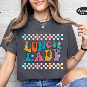 May include: A dark gray t-shirt with a checkered pattern and the text "Lunch Lady" in colorful, retro-style letters. The letters are outlined in black and have a white background.