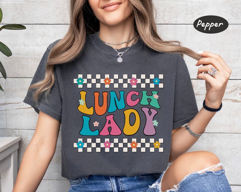 May include: A dark gray t-shirt with a checkered pattern and the text "Lunch Lady" in colorful, retro-style letters. The letters are outlined in black and have a white background.