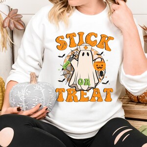 May include: White sweatshirt with a Halloween graphic. The design shows a ghost in a nurse's cap, holding a syringe and a jack-o'-lantern bucket. The words "STICK OR TREAT" are in orange, retro lettering.