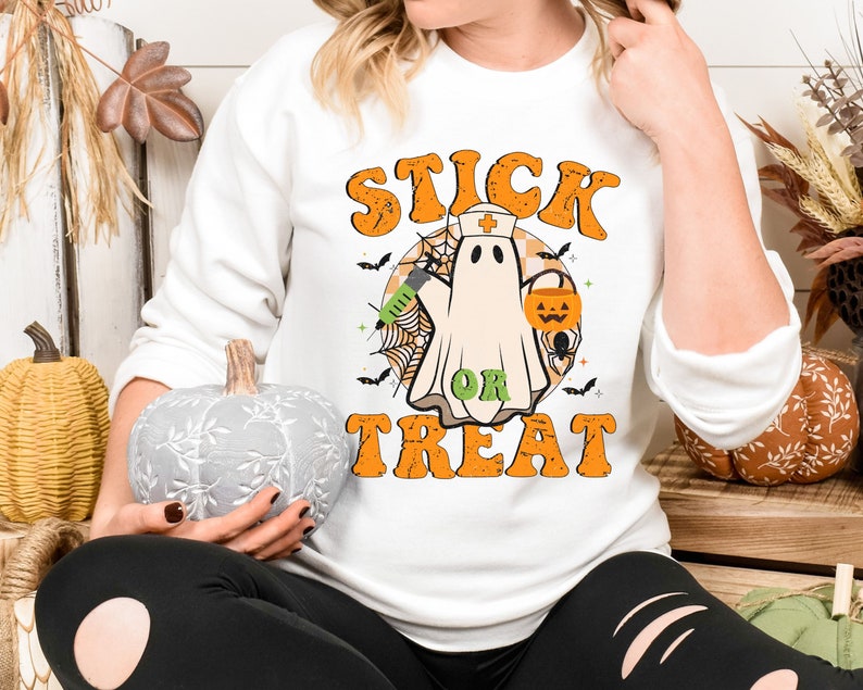 May include: White sweatshirt with a Halloween graphic. The design shows a ghost in a nurse's cap, holding a syringe and a jack-o'-lantern bucket. The words "STICK OR TREAT" are in orange, retro lettering.