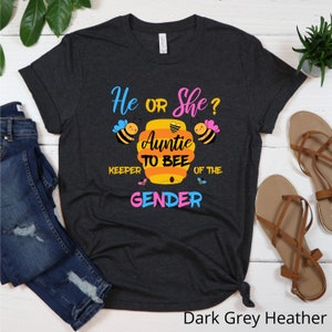 May include: Dark grey heather t-shirt with the text "He or She? Auntie to Bee Keeper of the Gender" in pink, blue, and yellow. The design features a beehive with a heart and bees.
