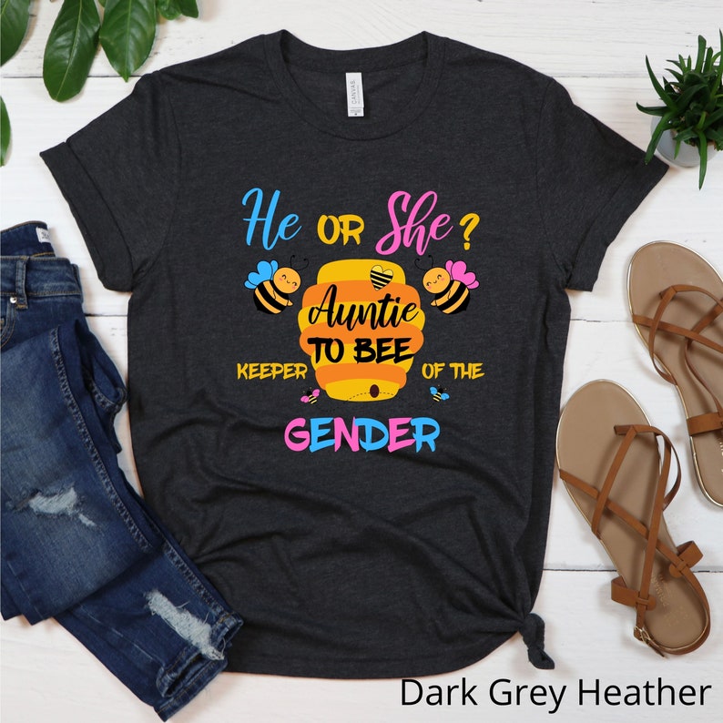 May include: Dark grey heather t-shirt with the text "He or She? Auntie to Bee Keeper of the Gender" in pink, blue, and yellow. The design features a beehive with a heart and bees.