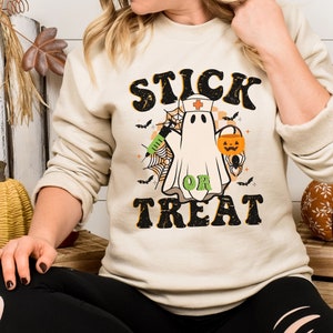 May include: Beige sweatshirt with a Halloween graphic. The design features a ghost in a nurse's cap, holding a syringe and a jack-o'-lantern bucket, with the words "STICK OR TREAT" in a retro font. Bats and spiderwebs complete the design.