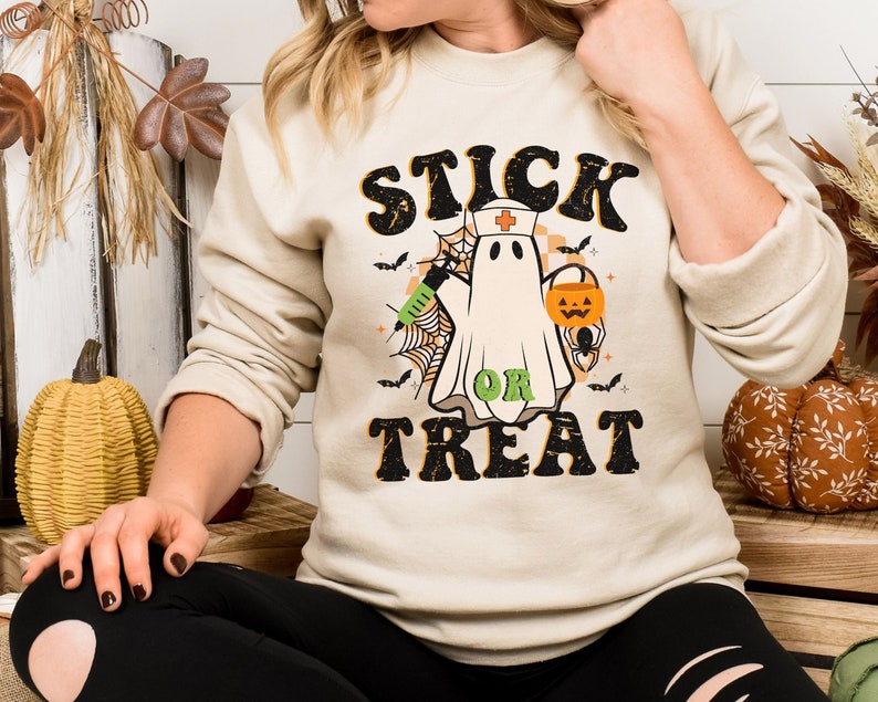 May include: Beige sweatshirt with a Halloween graphic. The design features a ghost in a nurse's cap, holding a syringe and a jack-o'-lantern bucket, with the words "STICK OR TREAT" in a retro font. Bats and spiderwebs complete the design.