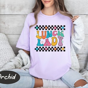 May include: A purple t-shirt with a checkered pattern and the text "Lunch Lady" in colorful letters. The letters are outlined in black and have a retro style.