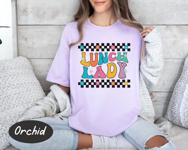 May include: A purple t-shirt with a checkered pattern and the text "Lunch Lady" in colorful letters. The letters are outlined in black and have a retro style.