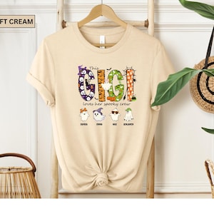 May include: A soft cream t-shirt featuring a graphic with the text This GIGI loves her spooky crew. The design includes four ghosts with names Sophia, Emma, Max, and Benjamin, decorated with Halloween motifs like pumpkins, bats, and spiders.