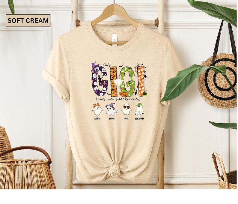 May include: A soft cream t-shirt featuring a graphic with the text This GIGI loves her spooky crew. The design includes four ghosts with names Sophia, Emma, Max, and Benjamin, decorated with Halloween motifs like pumpkins, bats, and spiders.
