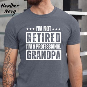 May include: Heather Navy t-shirt with the text "I'M NOT RETIRED I'M A PROFESSIONAL GRANDPA" in white block letters. The text is arranged in three lines with star accents. The shirt is a crew neck style.