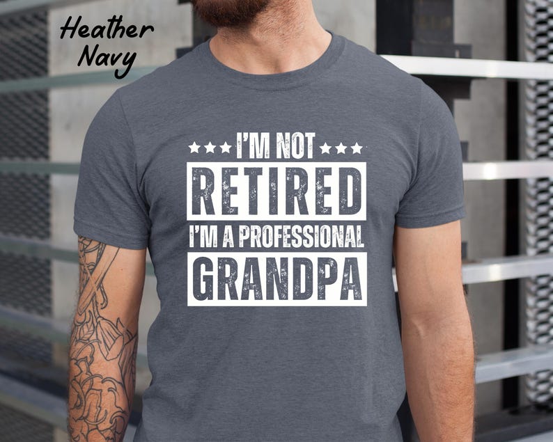May include: Heather Navy t-shirt with the text "I'M NOT RETIRED I'M A PROFESSIONAL GRANDPA" in white block letters. The text is arranged in three lines with star accents. The shirt is a crew neck style.