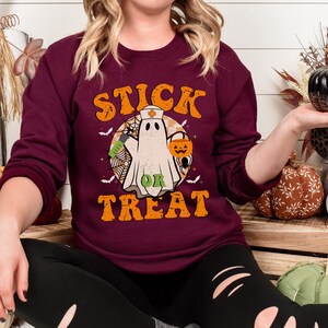May include: A maroon crew neck sweatshirt with the words "STICK OR TREAT" in orange lettering. The graphic features a ghost wearing a nurse's hat, holding a syringe, and a jack-o'-lantern. The design includes bats and a spider web, perfect for Halloween.