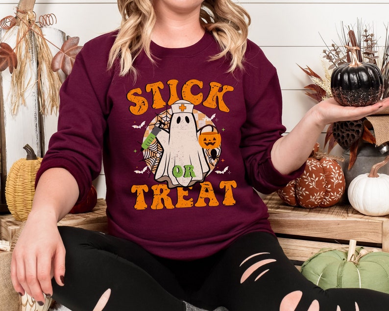 May include: A maroon crew neck sweatshirt with the words "STICK OR TREAT" in orange lettering. The graphic features a ghost wearing a nurse's hat, holding a syringe, and a jack-o'-lantern. The design includes bats and a spider web, perfect for Halloween.