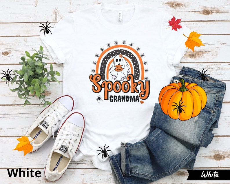 May include: White t-shirt with the text "Spooky Grandma" in orange and black, featuring a ghost holding a pumpkin. The design includes a rainbow and spiders, perfect for Halloween. The shirt is paired with jeans and white sneakers.