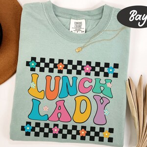 May include: A light blue t-shirt with a checkered pattern and the words "Lunch Lady" in colorful, retro-style lettering. The letters are outlined in black and have small flower accents.