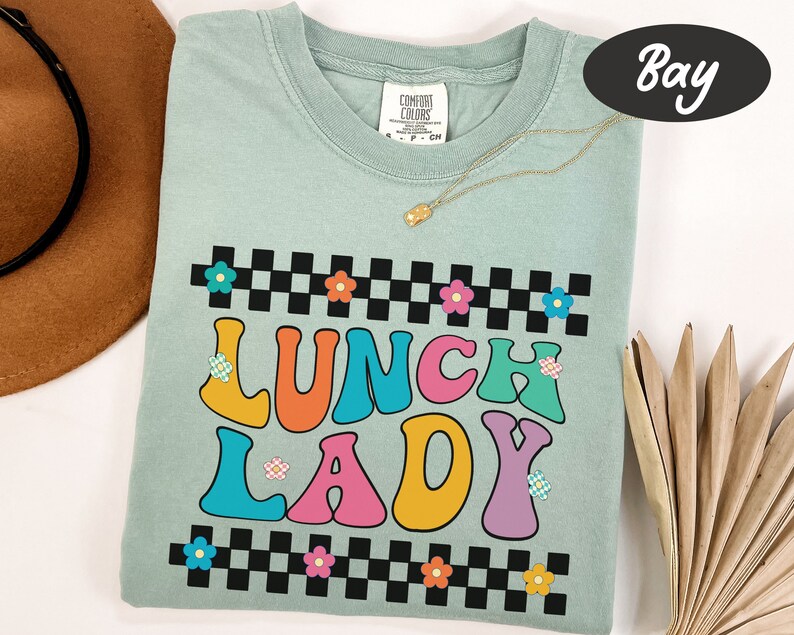May include: A light blue t-shirt with a checkered pattern and the words "Lunch Lady" in colorful, retro-style lettering. The letters are outlined in black and have small flower accents.