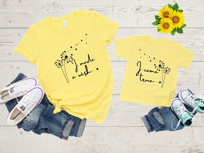 Op de afbeelding: Twee gele T-shirts met paardenbloemontwerpen en tekst. Het grotere shirt zegt "Ik heb een wens gedaan." Het kleinere shirt zegt "Ik ben uitgekomen."