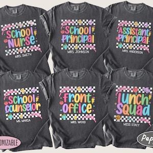 May include: Several dark gray t-shirts with colorful school-themed designs. Each shirt features a different title like "School Nurse" or "School Principal" with a name below. The designs include a checkered border and various school-related graphics.