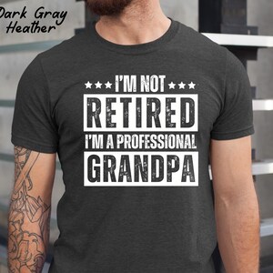 May include: Dark gray heather t-shirt with the text "I'M NOT RETIRED I'M A PROFESSIONAL GRANDPA" in white block letters. The shirt has a crew neck and short sleeves. The text is centered on the chest.