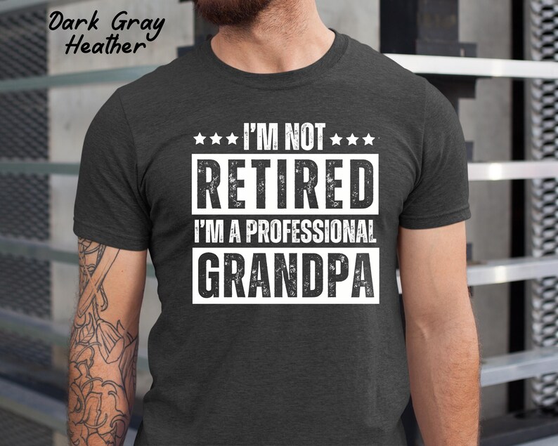 May include: Dark gray heather t-shirt with the text "I'M NOT RETIRED I'M A PROFESSIONAL GRANDPA" in white block letters. The shirt has a crew neck and short sleeves. The text is centered on the chest.