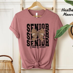 Puede incluir: Una camiseta de color malva jaspeado con un gr&aacute;fico negro que dice "Senior" tres veces con un lazo de estampado de leopardo en el medio. El texto "Class of 2026" est&aacute; debajo del gr&aacute;fico.
